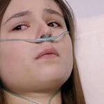 Clarinha deitada em uma cama de hospital, com os olhos abertos e um sorriso fraco no rosto. Álvaro, Dário e Flávia a observam com surpresa e emoção.