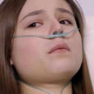 Clarinha deitada em uma cama de hospital, com os olhos abertos e um sorriso fraco no rosto. Álvaro, Dário e Flávia a observam com surpresa e emoção.