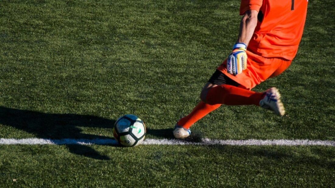 Goleiro de uniforme laranja batendo tiro de meta em campo de futebol com grama sintética