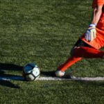Goleiro de uniforme laranja batendo tiro de meta em campo de futebol com grama sintética