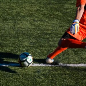 Goleiro de uniforme laranja batendo tiro de meta em campo de futebol com grama sintética